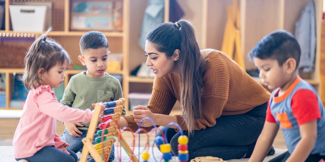 A female Kindergarten teacher of Middle Eastern decent, sits on the floor with students as they play with various toys and engage in different activities. They are each dressed casually as they learn through their play.