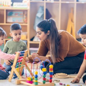 A female Kindergarten teacher of Middle Eastern decent, sits on the floor with students as they play with various toys and engage in different activities. They are each dressed casually as they learn through their play.