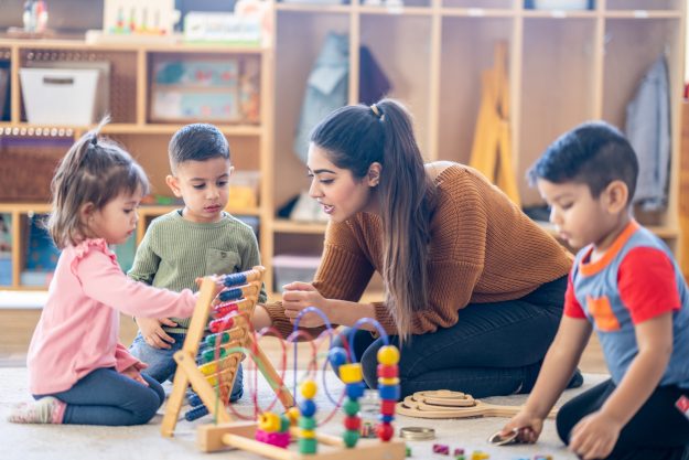 A female Kindergarten teacher of Middle Eastern decent, sits on the floor with students as they play with various toys and engage in different activities. They are each dressed casually as they learn through their play.